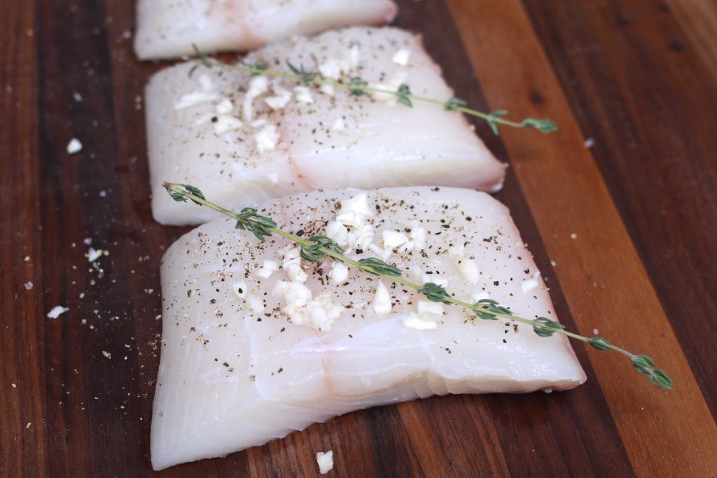 Fresh halibut filets seasoned with garlic, thyme, salt and pepper on a wooden cutting board.