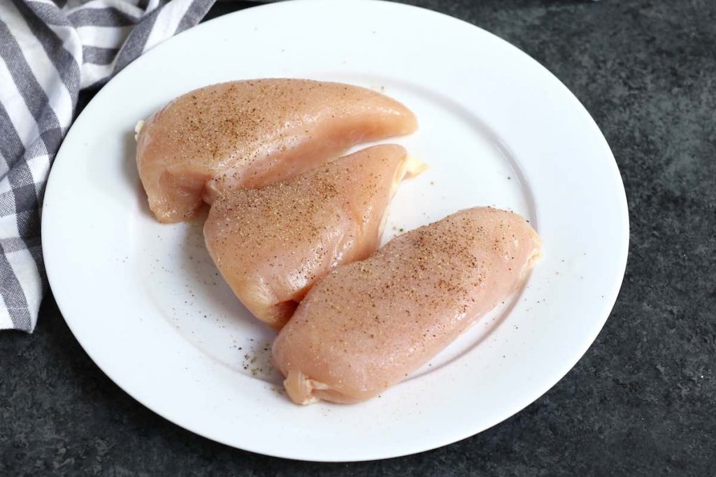 Raw chicken breasts seasoned with salt, pepper, and garlic powder on a white plate.