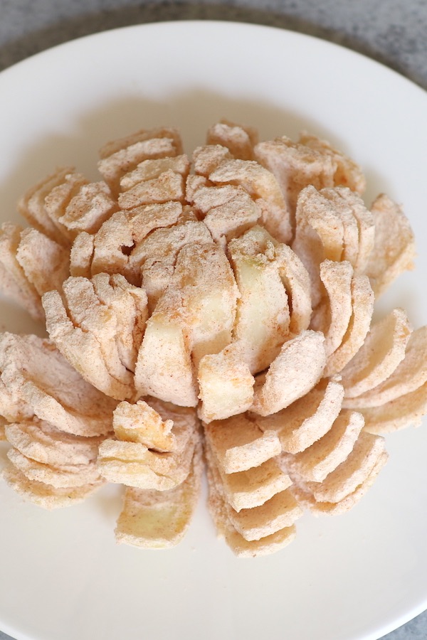 An onion cut to an onion flower, seasoned and battered. It’s placed on a white plate and ready to be deep fried.
