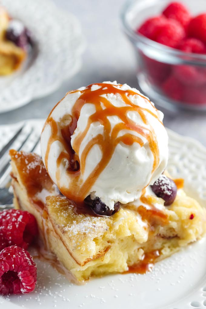 Baked blueberry French toast casserole served on a white plate with raspberries, dusted with powdered sugar, topped with vanilla ice cream and drizzled with caramel sauce!