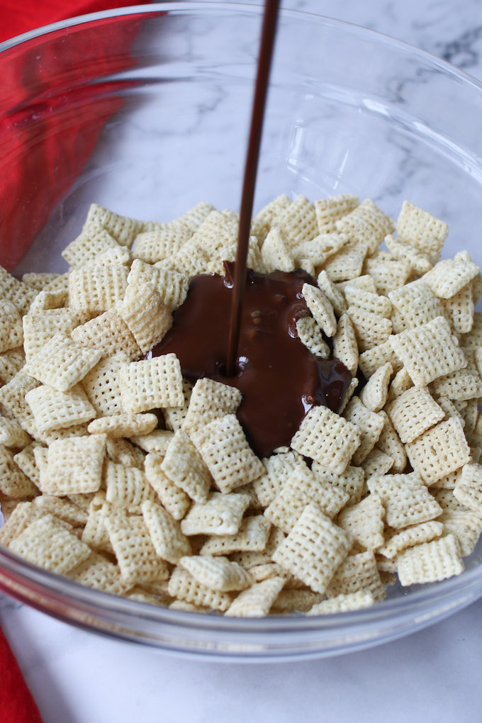 Adding chocolate and peanut butter mix into Chex cereal in a large clear bowl