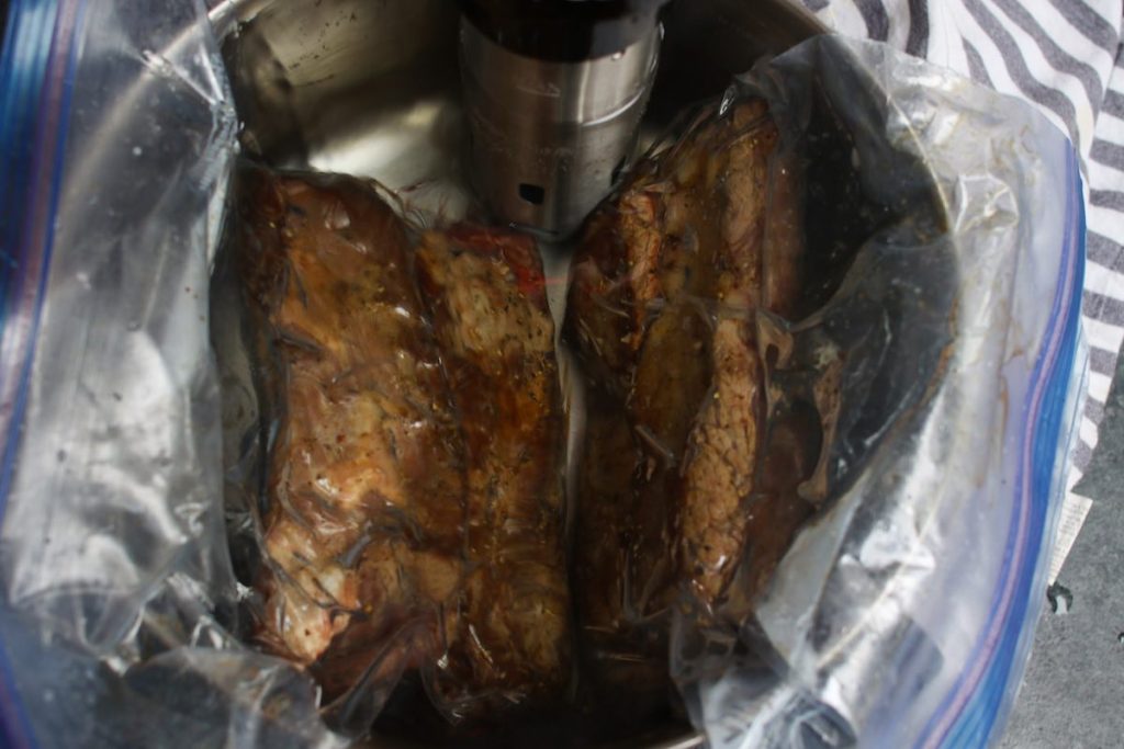 Closeup of two bags of vacuum-sealed short ribs cooking in sous vide warm water bath in a large pot.