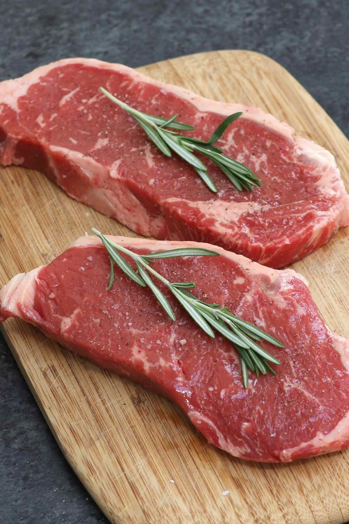 Raw New York strip steak seasoned with salt and pepper on a cutting board.