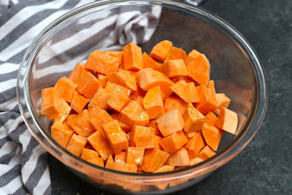 Sweet potatoes seasoned with oil, salt, and pepper in a large clear mixing bowl.
