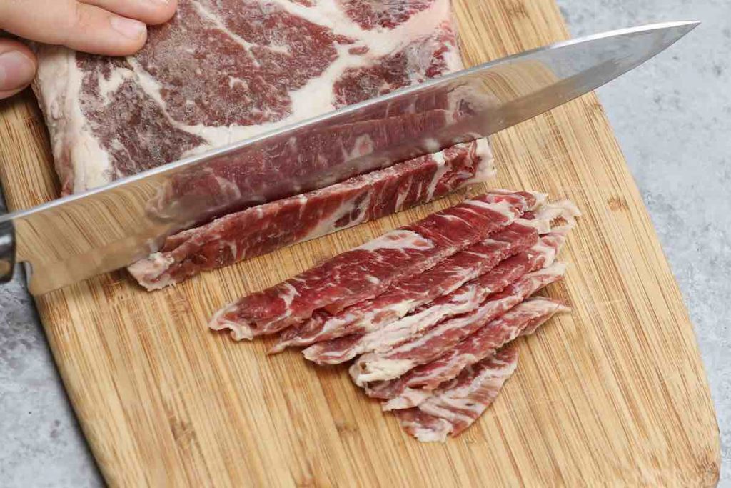 Cutting half-frozen beef into thin slices on a wooden cutting board.