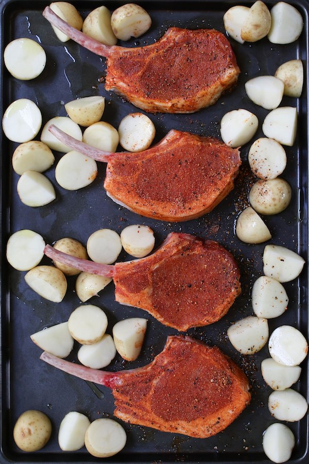 Four seasoned bone-in pork ribeye chops arranged on a baking sheet with halved potatoes