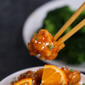Orange chicken piece held with chopsticks showing glossy sauce, sesame seeds, and green onions