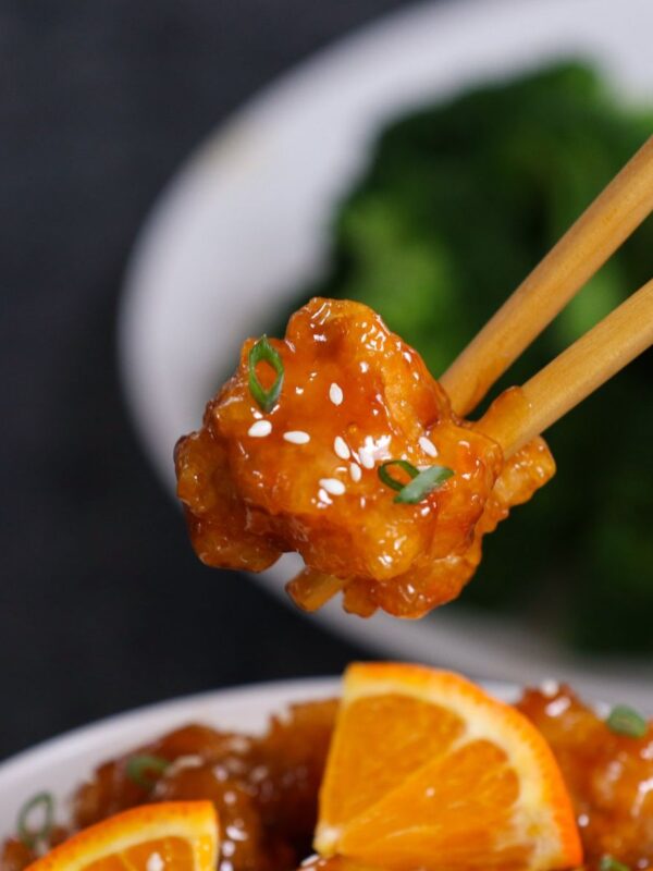 Orange chicken piece held with chopsticks showing glossy sauce, sesame seeds, and green onions