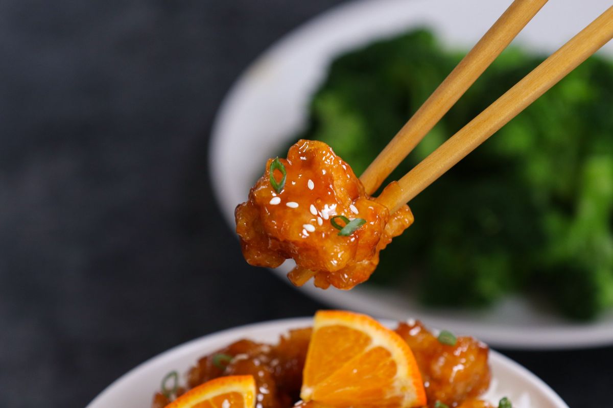 Orange chicken piece held with chopsticks showing glossy sauce, sesame seeds, and green onions