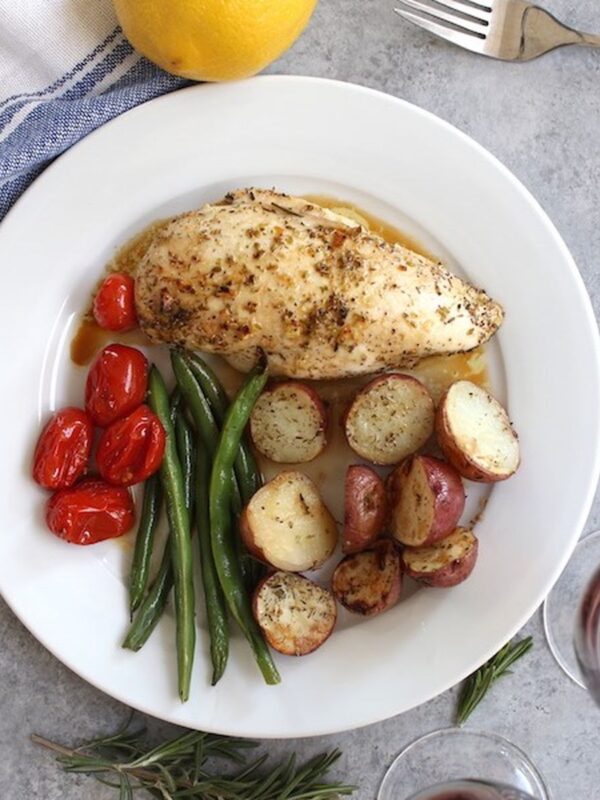 Baked chicken breast with roasted potatoes, green beans, and tomatoes on a plate with lemon, rosemary, and red wine
