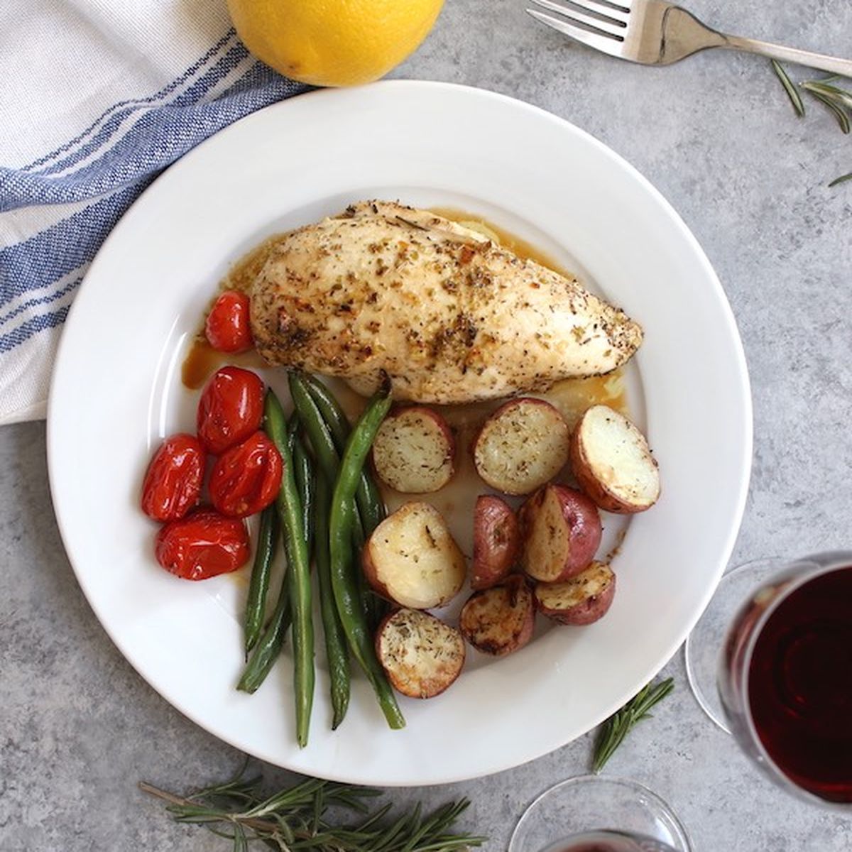 Baked chicken breast with roasted potatoes, green beans, and tomatoes on a plate with lemon, rosemary, and red wine