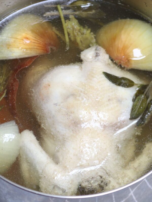 Whole chicken simmering in a pot with onions, herbs, and broth