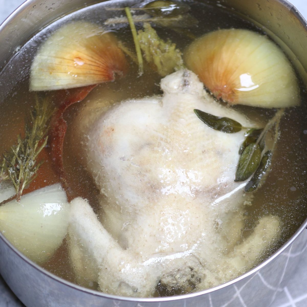 Whole chicken simmering in a pot with onions, herbs, and broth