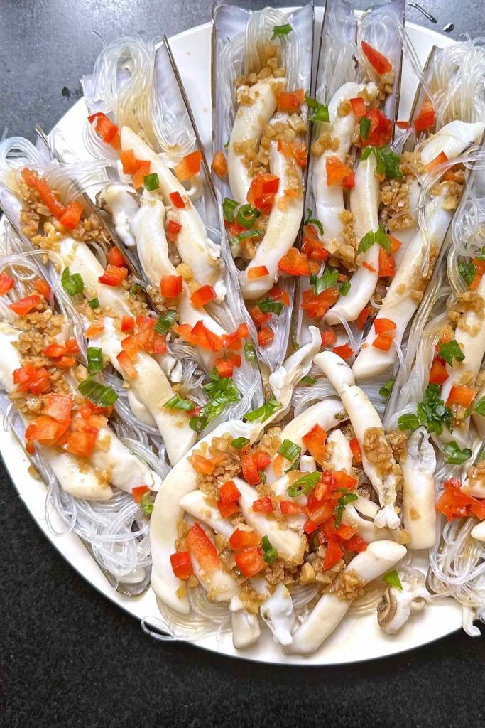 A plate with nicely arranged and cooked razor clams with garlic and rice noodles. Beautiful presentation with bright red and green colors.